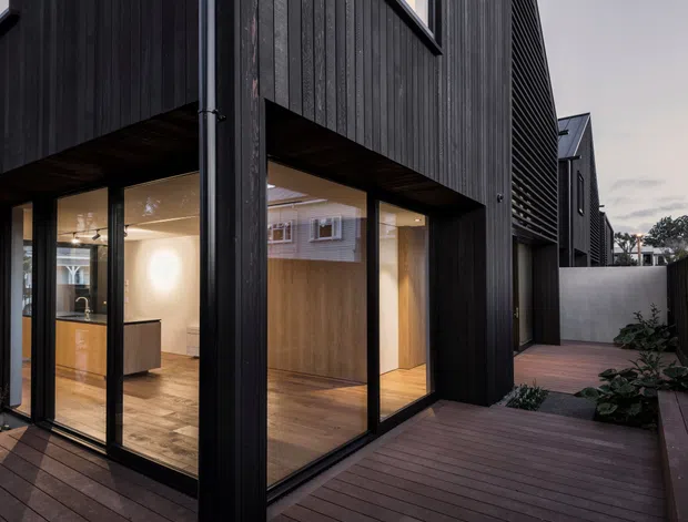 A contemporary multi-level residential exterior viewed from a timber deck or walkway. The facade is clad in dark vertical boards with projecting upper volumes, large black-framed floor-to-ceiling glazing, and a recessed warmly lit interior visible through the windows. A narrow landscaped side courtyard and timber privacy fencing run alongside the building.