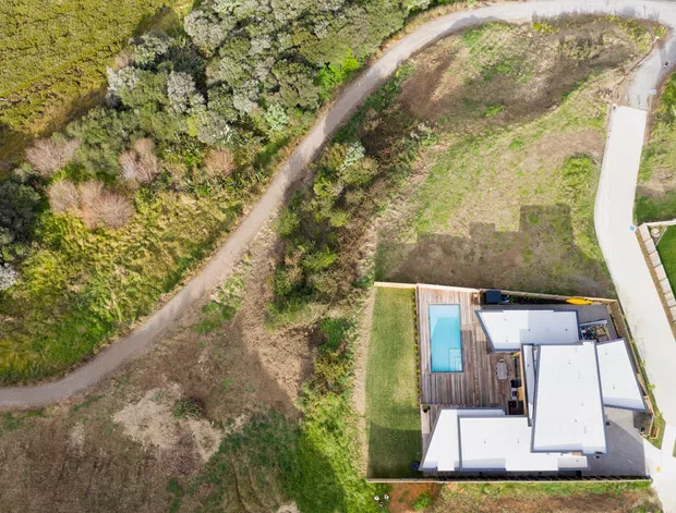 Aerial view of a contemporary residential property on a landscaped, semi-rural site. The home has several flat-roofed white volumes arranged around timber decking and a rectangular swimming pool, with curved access roads and extensive surrounding vegetation.