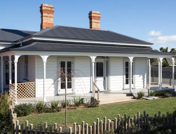 Street-facing exterior view of a restored single-storey Victorian-era cottage with white weatherboard walls, a broad corrugated metal hipped roof, two brick chimneys, and a covered front veranda supported by decorative timber posts and arches. The house has tall multi-pane doors and windows, latticework, a small landscaped front garden, and a low timber picket fence under clear daylight.