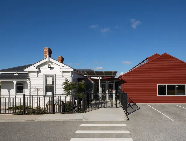 Exterior view of a renovated white weatherboard cottage adjoining a large red gabled building. The cottage has traditional detailing, a front veranda, chimneys and dark-framed windows, while the adjacent structure has simple contemporary cladding and a broad paved forecourt with pedestrian access.