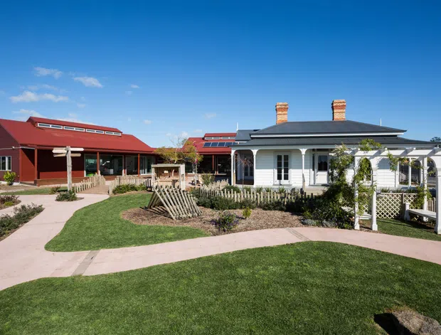 A sunny exterior view of a low-rise residential property comprising a white traditional cottage-like building with a dark hipped roof, brick chimneys, verandah and decorative white trellis, alongside a larger red-painted gabled building with roof skylights. Curving pale paved paths cross a landscaped lawn and garden beds. The scene has a heritage-inspired, country-style character with clear blue sky and open grounds.