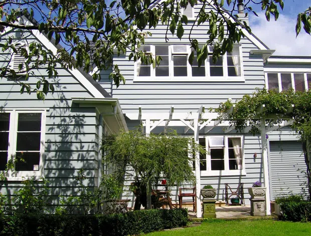 Exterior view of a light blue-grey weatherboard house with white trim, multi-pane windows, projecting gabled roof sections, and a leafy landscaped garden. A covered pergola and paved courtyard seating area sit against the facade, with mature trees framing the view.