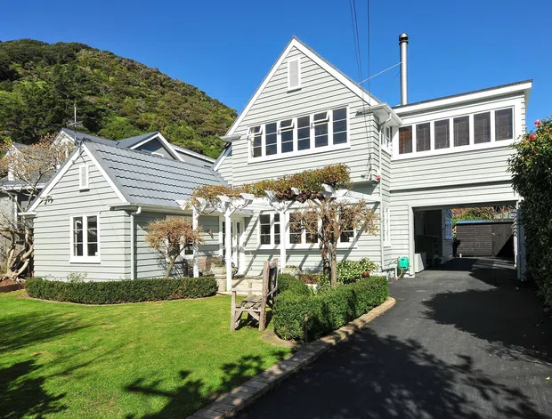 A bright exterior view of a multi-wing, multi-storey house with pale grey-green horizontal weatherboard cladding, steep gabled roofs, white-trimmed windows, and a covered driveway or garage passage. The home sits beside a steep vegetated hillside, with a manicured lawn, clipped hedges, and established garden planting in the foreground.