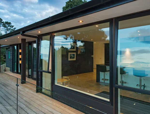 Exterior view of a contemporary, single-storey house with dark horizontal cladding, broad floor-to-ceiling glazing, and a low-pitched roof. A timber deck with frameless glass balustrading runs along the facade, while the interior and ocean beyond are visible through the windows.