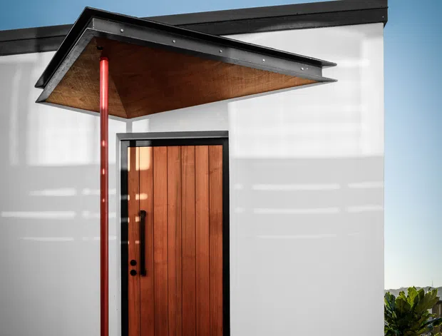Close-up exterior view of a minimalist modern building entrance. A warm vertical timber door with dark metal hardware is set into a crisp white facade, sheltered by a dramatic projecting triangular canopy with a timber soffit and black metal edging. A slender red support post adds a bold accent. The entrance sits on a dark textured paved surface, with clear blue sky and partial planting visible at the edges.