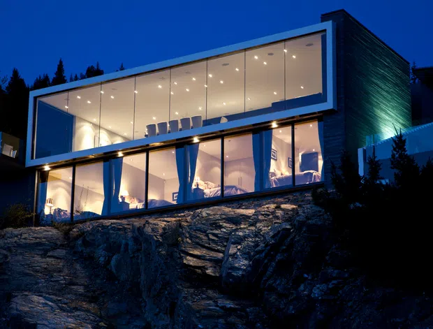 A modern multi-storey house dramatically positioned on a rocky, sloped site, photographed at dusk. The building features a cantilevered rectangular upper volume, extensive floor-to-ceiling glazing, dark horizontal cladding, and warm illuminated interiors visible through the glass.