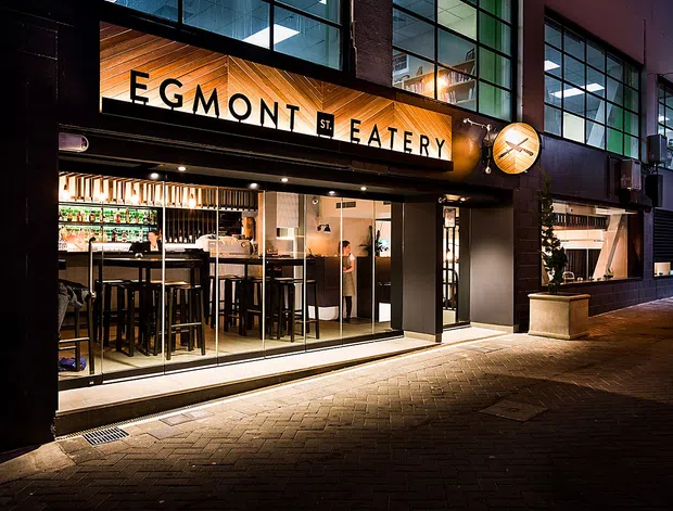 Night-time exterior view of the Egmont Street Eatery, a contemporary urban restaurant and bar front. The dark facade features extensive glazed shopfront windows and doors, warm timber soffit and signage, illuminated interior bar seating, and a paved pedestrian frontage. The building is integrated into a multi-storey commercial facade with large upper-level windows.