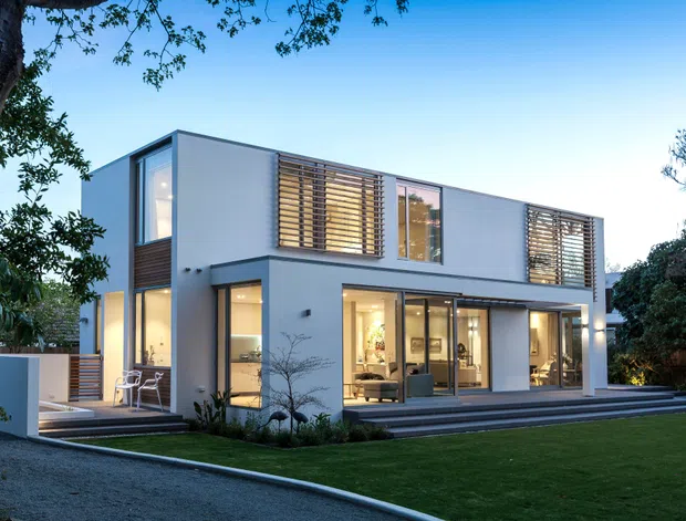 Modern two-storey residential house viewed from the landscaped front or rear garden. The building has clean white rendered walls, extensive floor-to-ceiling glazing, dark metal-framed windows and doors, and horizontal timber louvres screening upper-level windows. Broad steps lead to a glazed living area and a neatly planted lawn, with mature trees surrounding the property.