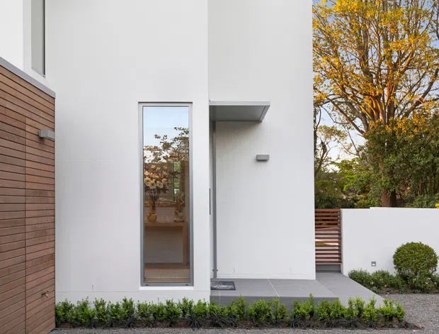A clean contemporary residential facade viewed from the front, featuring smooth white rendered walls, warm horizontal timber cladding, a narrow full-height glazed entry door, a simple projecting canopy, and a grey paved entry terrace. A low white boundary wall, timber slat gate, clipped planting, and mature deciduous trees frame the entrance.