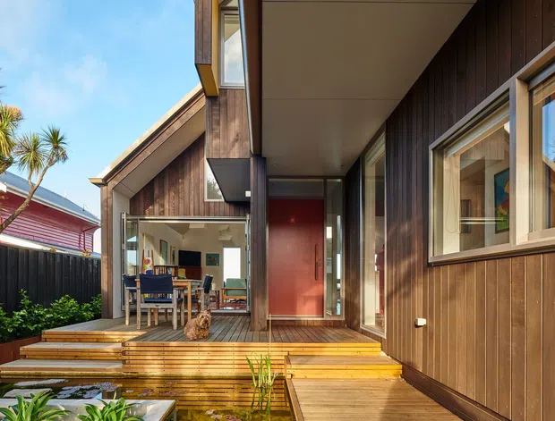 Exterior view of a contemporary timber-clad home entry and raised deck. The image shows a covered walkway leading to a red front door, warm timber steps and decking, large windows, and a partially visible dining area beyond the glazing. The building sits in a landscaped suburban setting with distinctive spiky ornamental trees and a neighbouring structure visible in the background.