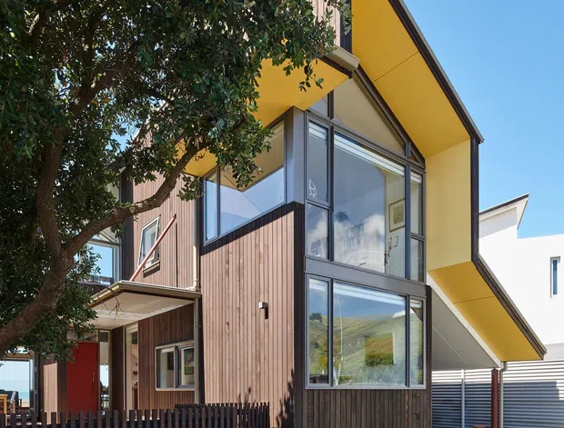 Exterior architectural photograph of a contemporary multi-level home with a dramatic yellow sloping roof canopy, dark timber vertical cladding, extensive glazed corner windows, and metal-framed glazing. A landscaped front area with mature trees, dark picket fencing, paving, and ornamental planting occupies the foreground.