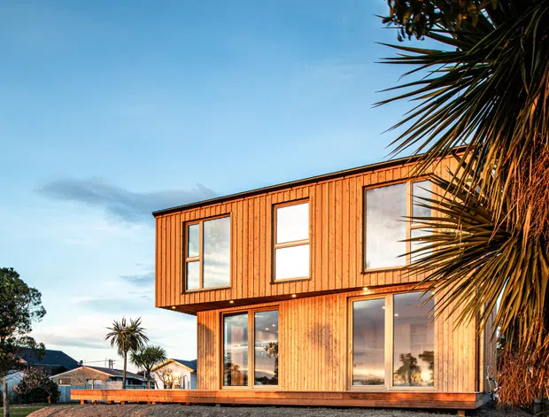 A modern two-storey timber-clad house viewed from the exterior at sunset. The box-like upper level projects slightly over the lower floor, with extensive floor-to-ceiling glazing and vertically oriented windows. A timber deck runs along the ground floor, while mature flax and palm-like planting frame the right side. The warm wood facade contrasts with the blue sky and suburban coastal-looking setting.