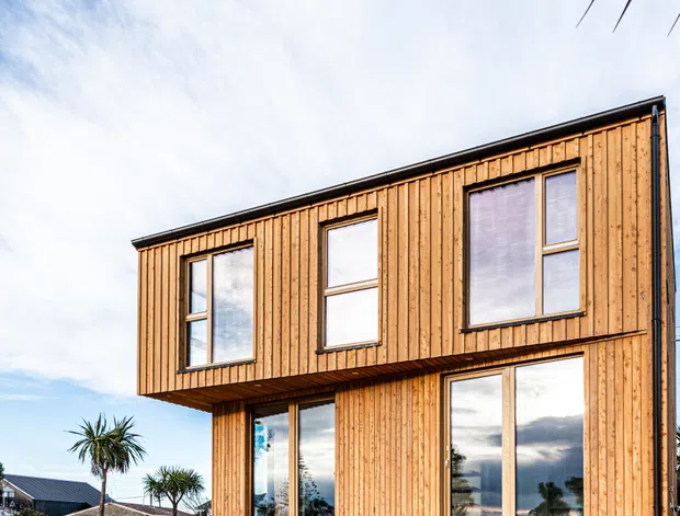 Exterior view of a contemporary two-storey rectangular house clad in warm vertical timber boards. The facade has large, vertically proportioned windows, a projecting upper floor, and a simple dark roof edge against a bright sky. Neighbouring homes and palm-like planting are visible in the background.