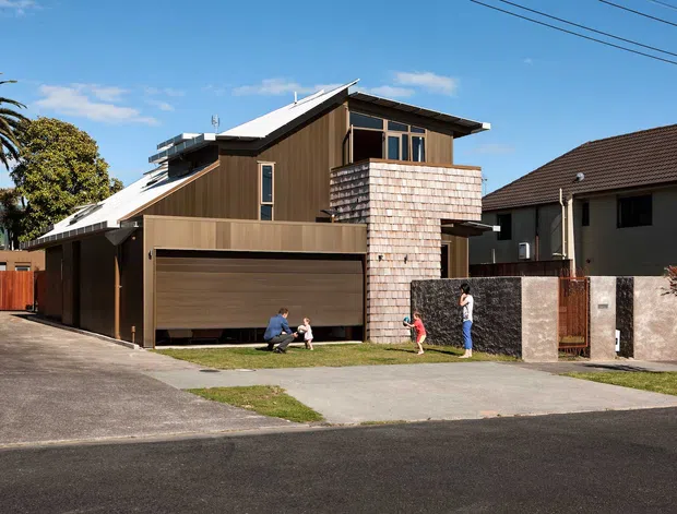 Street-facing exterior view of a contemporary multi-level residence with a sharply pitched, asymmetrical roof, dark vertical timber-like cladding, a large integrated garage door, and a projecting upper-level balcony or bay clad in light shingle-like panels. The house sits among neighbouring suburban homes, with a driveway, front lawn, boundary wall and fence visible. Several people are present outside, but the building is the main subject.