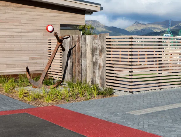 Exterior view of a contemporary low-profile building beside a paved driveway or walkway. The facade combines horizontal light timber cladding with a dark roof edge, while a weathered timber privacy screen and slatted timber fence enclose a small landscaped area. A large rusted anchor forms a decorative feature near the entrance. Mountains and ski-lift infrastructure are visible in the background.