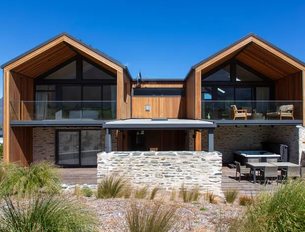 Exterior view of a contemporary alpine-style residence with two prominent timber-clad gabled wings, large dark-framed glazing, balconies, and a central recessed entrance or connecting volume. The lower level combines dark stone masonry with timber, while a low stone wall and outdoor seating occupy the foreground. Native grasses, gravel landscaping, and surrounding mountain terrain reinforce the rugged mountain setting.