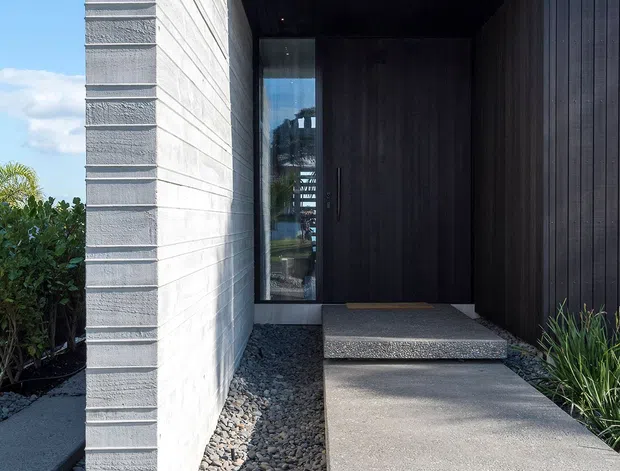 Contemporary residential entrance with a minimalist composition. A dark vertically clad facade frames a recessed timber-toned front door beneath a deep covered entry, contrasting with a pale textured masonry wall. Large rectangular concrete stepping slabs form the walkway through a bed of grey gravel, with sparse architectural planting along the edges.