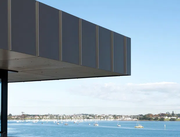 Exterior architectural view of a contemporary waterfront building with a dramatic cantilevered upper volume clad in dark vertical panels. A glass corner and exposed concrete soffit frame a broad harbour scene with moored sailboats and residential shoreline beyond.