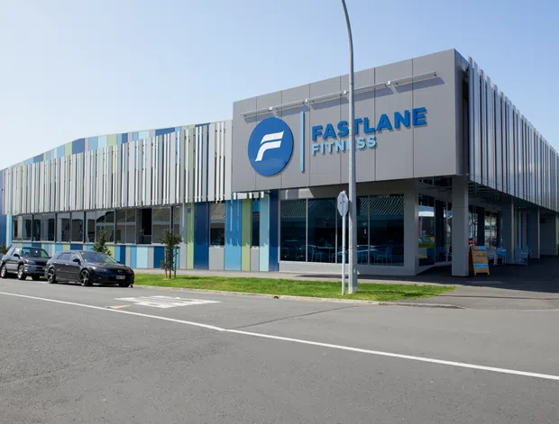 Street-level exterior view of a modern commercial fitness centre. The building has a long rectilinear form, a projecting upper volume, extensive glazing at the entrance, and a facade composed of closely spaced vertical fins in varied blue, grey, and white tones. Large blue fitness-centre signage is mounted prominently on the front.
