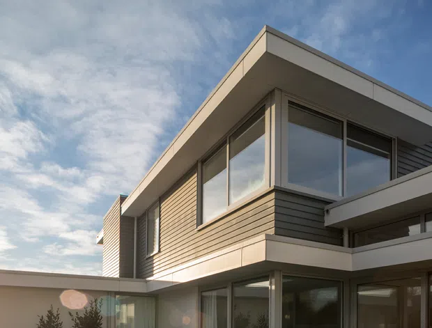 Exterior architectural photograph of a contemporary two-storey residence with a flat roof, strong horizontal lines, extensive glazing, pale rendered or metal fascia, and horizontal timber-look wall cladding. The low-angle view emphasizes the projecting upper-storey volume and sheltered ground-floor outdoor area.