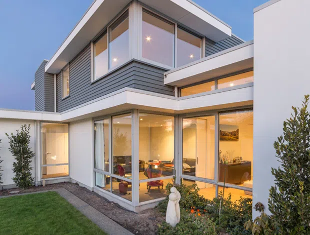 Exterior view of a contemporary multi-level home at dusk, featuring crisp white rendered volumes, grey horizontal cladding, broad roof overhangs, and extensive floor-to-ceiling glazing. Warm interior lighting reveals living areas and a kitchen through the windows. The house opens onto a landscaped courtyard-style garden with lawn, planted beds, and a narrow paved path.