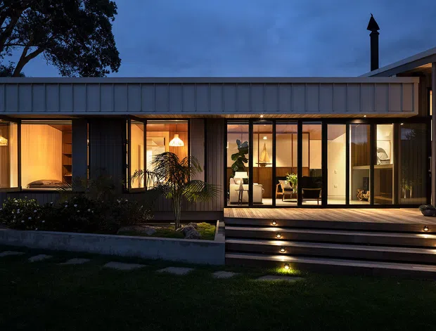 Twilight exterior view of a low, single-storey contemporary home with dark vertical cladding, broad horizontal roof forms, extensive glazed doors and windows, and a warmly illuminated interior. A raised timber deck and stepped entry with integrated lighting lead through landscaped garden beds to the house.