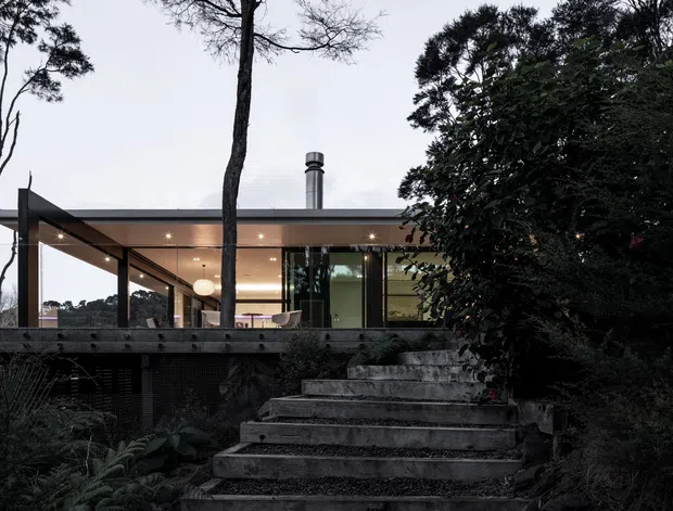 Exterior view of a contemporary elevated pavilion-style house set among mature trees and dense garden planting. A broad covered terrace with slim dark structural posts, warm recessed lighting, extensive glazing, and a visible interior fireplace opens onto a timber deck. Broad concrete steps lead up through the landscaped garden to the terrace.