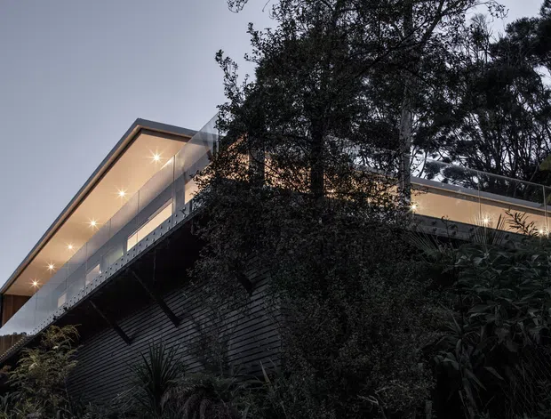 Dusk exterior view of a contemporary multi-level residence set into a densely landscaped, sloping site. The building features a long projecting upper volume with extensive glazing, illuminated recessed soffit lighting, slim dark roof planes, and dark horizontal cladding over a textured retaining wall.