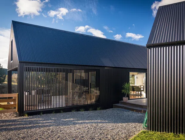 Exterior view of a contemporary dark-clad building composed of steeply pitched gabled volumes. Vertical ribbed metal cladding covers the walls and roofs, with a screened outdoor area, gravel courtyard, timber fencing, and a glazed entrance revealing a dining space inside.