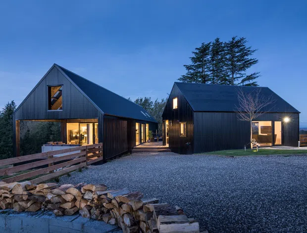 Twilight exterior view of two dark-clad gabled buildings arranged around a gravel forecourt. The buildings feature steep pitched roofs, vertical dark metal or timber-like cladding, warm illuminated windows, and timber fencing with stacked firewood in the foreground. A wooded landscape and distant water or horizon are visible beyond the site.
