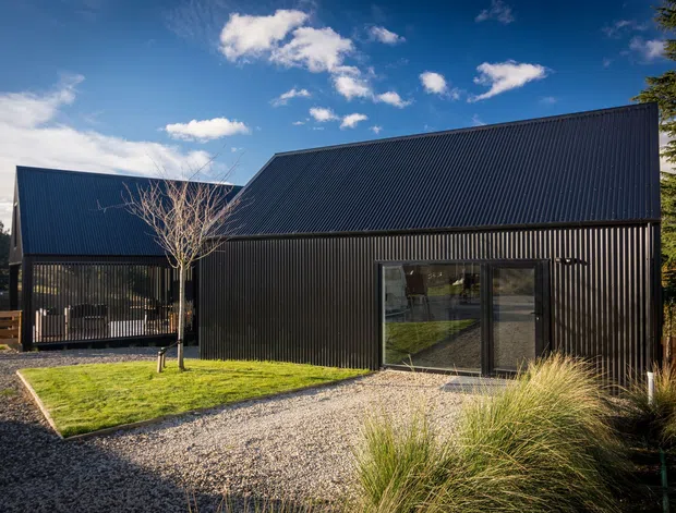 Exterior view of a contemporary dark-clad building with steep pitched roof forms, large glazed doors, gravel landscaping, ornamental grasses, timber fencing, and mature trees under a clear sky.
