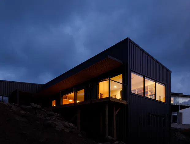 A modern dark-clad house photographed at dusk on a rocky sloping site. The angular, gabled volume features vertically profiled black cladding, extensive warm-lit glazing, a projecting covered terrace or balcony, and timber-lined soffits supported above the rocky ground. The dramatic blue evening sky contrasts with the warm interior lighting.