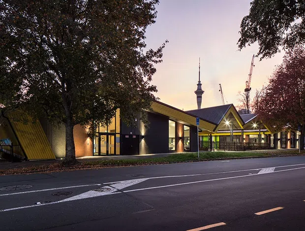 Twilight exterior view of a contemporary low-rise civic or commercial building set along an urban street. The building features angular monopitched and gabled roof forms, dark facade panels, extensive glazing, warm linear lighting, and a covered entry or walkway. Mature street trees frame the scene, while a tower and construction cranes are visible in the background.