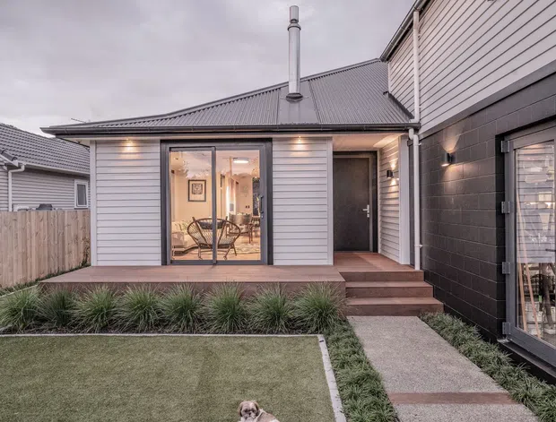Exterior view of a compact contemporary residential extension or rear courtyard, featuring light horizontal weatherboard cladding, a dark masonry-clad volume, a pitched metal roof with chimney, glazed doors opening to a timber deck, and a landscaped lawn with ornamental grasses.