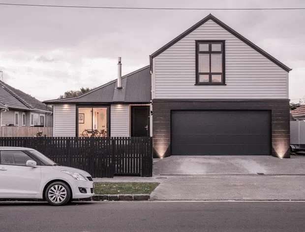 Street-facing view of a compact two-storey residential home with a steep front gable, light horizontal weatherboard cladding, dark brick or masonry at ground level, a large integrated garage, and a dark metal pitched roof. A fenced front yard, driveway, neighbouring homes, and a parked car are visible.