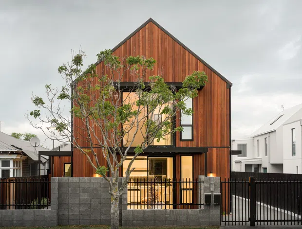 Front exterior view of a narrow multi-storey residence with a steep gable roof and vertically clad warm timber facade. Large windows and a recessed balcony/opening face the street, with a dark metal fence, concrete block boundary wall, mature tree, and neighbouring urban homes visible.