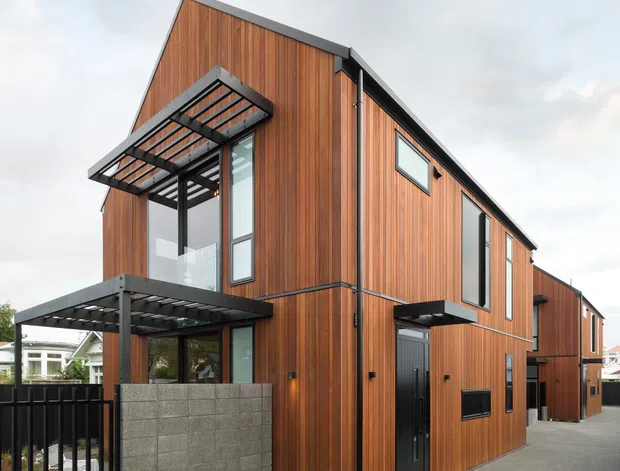 Exterior view of a contemporary multi-storey townhouse or compact residential development, clad predominantly in warm vertical timber with dark metal window and door frames. The building features sharp gabled roof forms, projecting metal-framed balconies or canopies, extensive glazing, and a rendered masonry boundary wall. It is photographed from a paved driveway or access lane.