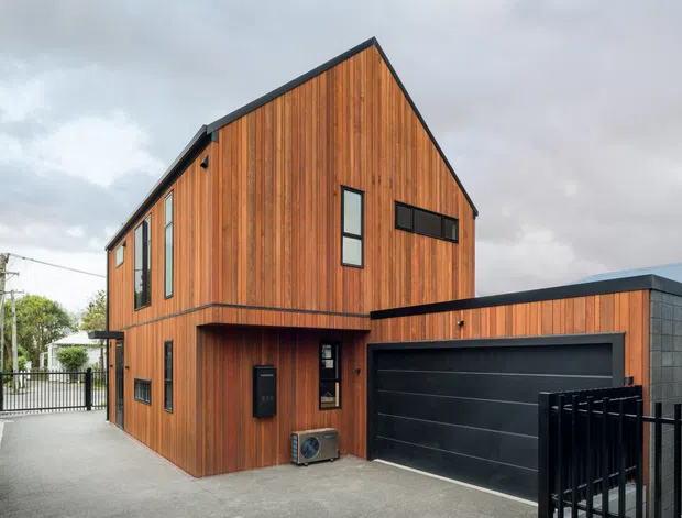 Exterior view of a contemporary multi-level residence with a steep gable roof and vertical warm timber cladding. The home includes a projecting upper storey, narrow dark-framed windows, a large black sectional garage door, and a lower flat-roof garage volume with dark masonry or blockwork. A paved driveway and black metal fencing frame the foreground.