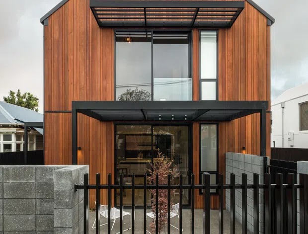 Front exterior view of a compact two-storey gabled house with warm vertical timber cladding, dark metal roof edging, large glazed openings, projecting black metal awnings, a black metal pedestrian fence, and grey concrete block boundary walls.