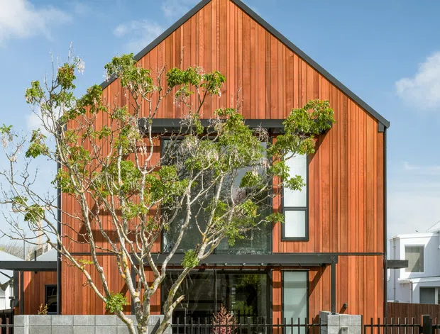 Street-facing view of a contemporary two-storey residence with a steep gable roof and prominent vertical warm timber cladding. Large glazed openings sit behind a low grey concrete-block wall and black metal fencing, with a paved entry path and established street tree in the foreground.
