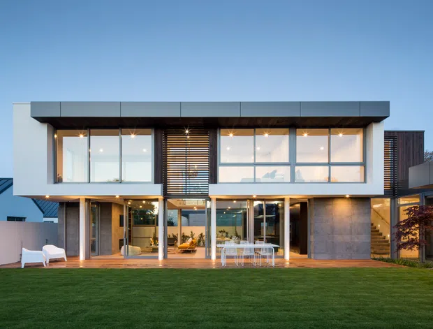 A contemporary two-storey residence viewed from the rear garden at dusk. The façade features broad floor-to-ceiling glazing, crisp white rendered volumes, dark metal roof and trim, timber screening, exposed structural posts, and a covered outdoor terrace with dining furniture. A large lawn occupies the foreground, with warm interior and exterior lighting creating an inviting connection between the house and garden.