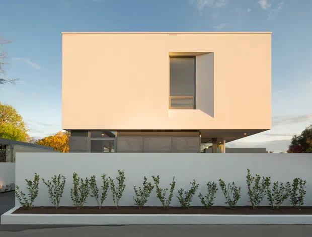 Front exterior view of a minimalist two-storey residence with a clean rectangular upper volume, pale peach rendered facade, deeply recessed vertical window, and a darker glazed lower level partly screened by a white boundary wall and linear planted garden bed. The house has a flat roof and strong cantilevered overhangs, set against a clear sky and mature landscaping.