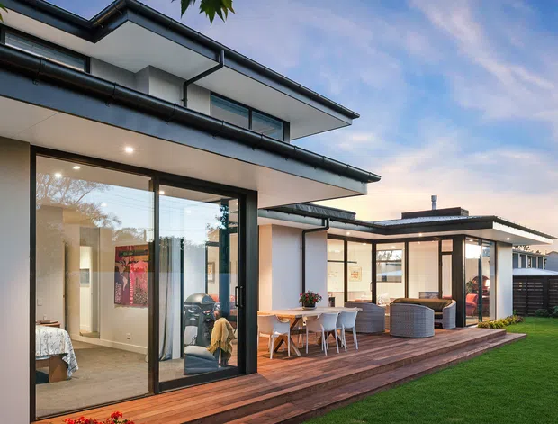 Contemporary multi-level home viewed from the garden, with crisp white rendered walls, dark low-pitched roof forms, extensive black-framed glazing, and a timber deck extending into a covered outdoor dining and lounge area. The image also shows a neat lawn and partial interior spaces through the glass.