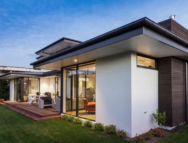 Exterior view of a contemporary multi-level residence with a low-pitched flat roof, strong horizontal overhangs, white rendered walls, dark timber-look vertical cladding, and extensive floor-to-ceiling glazing. A covered outdoor terrace with seating connects to the living spaces, surrounded by a manicured lawn and planted garden beds.