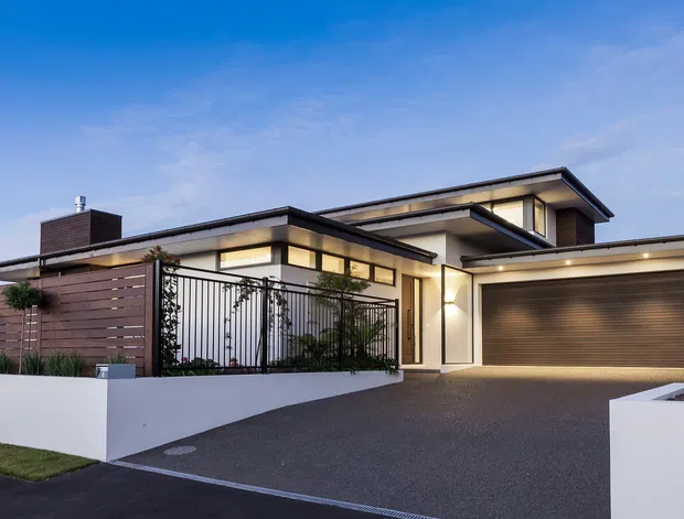 Contemporary multi-level residence viewed from the street at dusk, featuring clean horizontal rooflines, pale rendered walls, dark trim, extensive glazing, a timber privacy fence, a metal pedestrian gate, landscaped raised planters, and a wide integrated double garage with a dark roller door. The home is approached by a broad dark paved driveway.