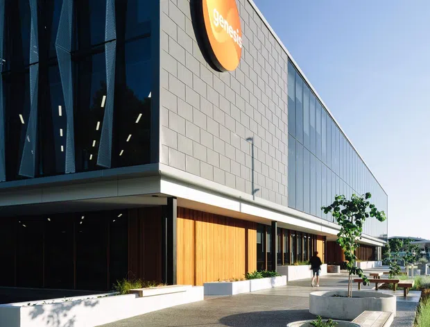 Exterior view of a contemporary commercial building with a strong geometric composition. The facade combines dark glazed curtain walling with deep vertical fins, pale rectangular masonry or tile cladding, warm timber wall panels, and a long glazed side elevation. A prominent circular orange sign is mounted high on the wall. The landscaped forecourt includes concrete planters, bench seating, paved walkways, and low planting.