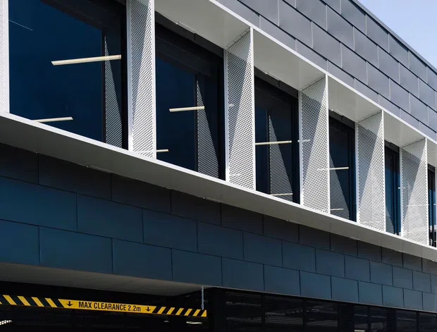 Contemporary multi-storey commercial building facade viewed from outside, featuring dark blue-grey metal cladding, tall glazed windows framed by white perforated metal screens, and a secure ground-level car park entrance with black mesh gates and a yellow clearance sign.