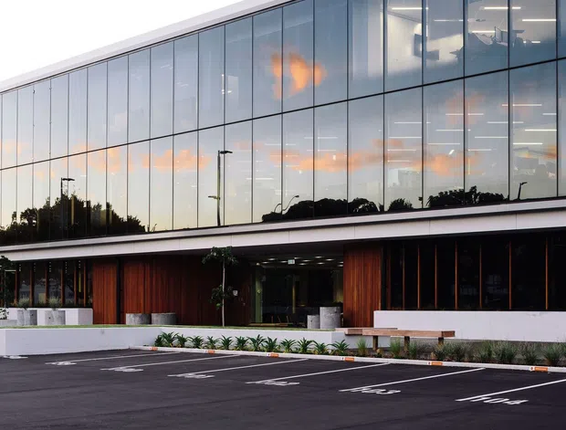 Exterior view of a contemporary multi-storey commercial building with a long glazed curtain wall, white horizontal slab edges, and warm vertical timber-clad sections at ground level. The building fronts a freshly marked asphalt parking area with landscaped planting along the entrance.