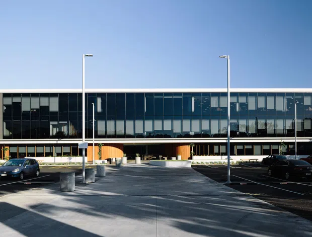 Wide exterior view of a large contemporary multi-storey commercial building with a long glazed curtain-wall facade, dark vertical framing, pale masonry side walls, and a covered ground-level entrance. The building fronts a paved forecourt and parking area with tall minimalist light poles under a clear blue sky.