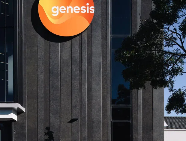 Exterior view of a contemporary commercial building with a tall, narrow facade clad in dark vertically ribbed panels. A prominent circular orange Genesis sign is mounted high on the wall, beside a slim vertical strip of dark glazing. The foreground includes landscaping, a bench, fencing, and a partial adjacent glazed volume. Strong sunlight creates crisp shadows against a clear blue sky.